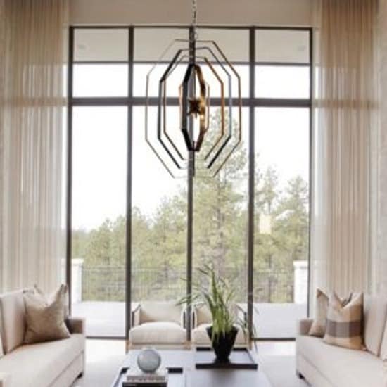 Modern living room featuring two beige sofas, an Orbita Series geometric chandelier, a glass coffee table with a plant, and expansive floor-to-ceiling windows with views of trees.