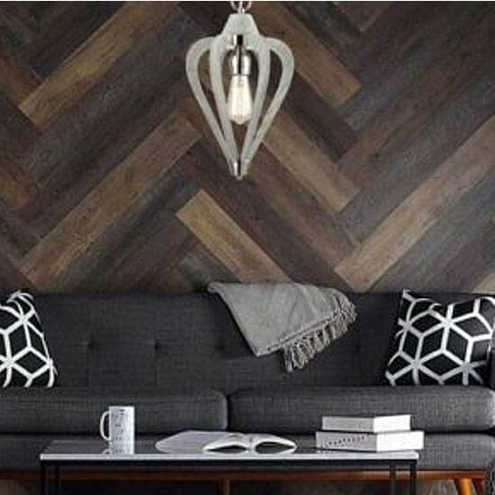 A modern living room showcases the Senorita Series: a dark gray sofa with geometric pillows, a cozy gray throw, and a wooden coffee table holding books and a mug, set before a herringbone wood wall under a chic pendant light.