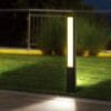 A Hathor series LED Bollard brightens a grassy area by the sidewalk, with metal railings and shrubs in the background.