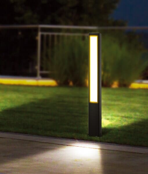 A Hathor series LED Bollard brightens a grassy area by the sidewalk, with metal railings and shrubs in the background.