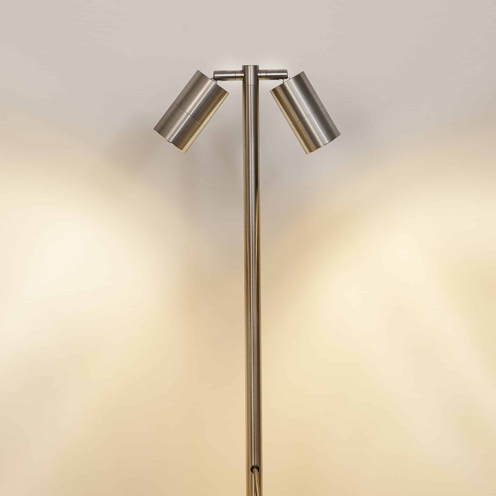 The Tivah HV1405-SS316 is a metal floor lamp with two cylindrical shades angled apart, casting light on a plain wall—ideal for achieving a Spike Spotlight effect in any modern space.