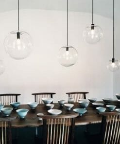 A wooden dining table set with ceramic bowls and plates, surrounded by wooden chairs, is illuminated by a row of Linder Selene Replica Clear Pendant lights.