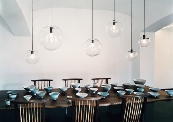 A wooden dining table set with ceramic bowls and plates, surrounded by wooden chairs, is illuminated by a row of Linder Selene Replica Clear Pendant lights.