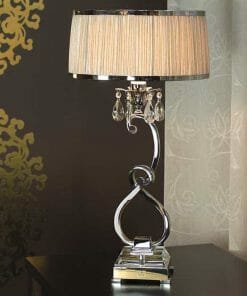 The Luxuria 1 Light Table Lamp showcases a metallic twisted base, crystal accents, and a beige pleated shade, stylishly placed on a dark surface by a patterned wall and sheer curtain.