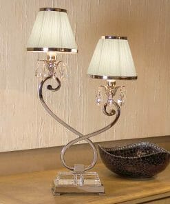 The Luxuria 2 light table lamp, featuring a double-shaded metal scroll design and crystal accents, is displayed on a wooden surface beside a dark decorative bowl.