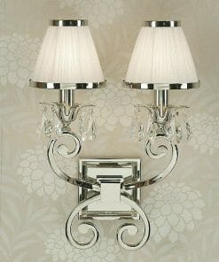 The Luxuria 2 light wall features dual arms with silver metalwork, crystal accents, and white pleated lampshades, set against floral-patterned wallpaper.