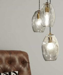 Three Organic large pendants with visible bulbs hang above the corner of a brown tufted leather chair placed against a light gray wall.