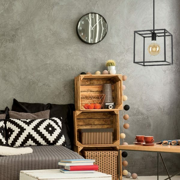 Modern living room with a gray textured wall, wooden crate shelves, black and white pillow, wall clock, Collins 6 light pendant, and two red cups on a small table.