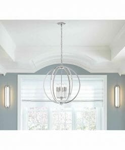 A Corinne 3 small pendant chandelier with a modern silver finish and spherical frame hangs from a white ceiling in a bright room featuring two wall sconces and large windows.
