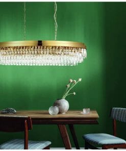 The Valparaiso 9G pendant, featuring an oval design in gold and crystal, is suspended above a wooden dining table with modern chairs, complemented by a green wall backdrop.