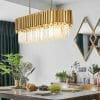 A contemporary dining room showcases the Regina Brass bar chandelier over a set table with fruit, plants, and geometric wall mirrors in the background.