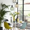 Modern dining area with a round white table from the Maracan range, vibrant chairs, pendant lights, greenery, and decorative vases by large windows overlooking a garden.