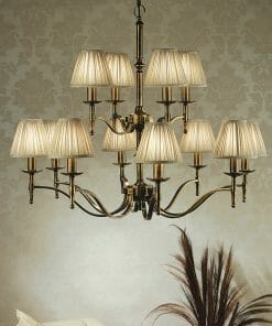 The Standford 3 Antique brass Shimmer shade chandelier with twelve arms and pleated beige shades hangs from the ceiling against patterned wallpaper.