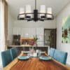 A modern dining room featuring a wooden table for four with teal chairs, a floral centerpiece, and the Devon 6 light Black chandelier; kitchen area visible in the background.
