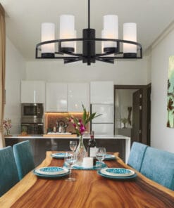 A modern dining room featuring a wooden table for four with teal chairs, a floral centerpiece, and the Devon 6 light Black chandelier; kitchen area visible in the background.