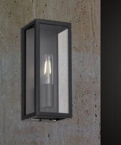 The Arazano 25 Solid Brass Wall Light features a rectangular black metal frame and clear glass panels, mounted stylishly on a textured stone wall.