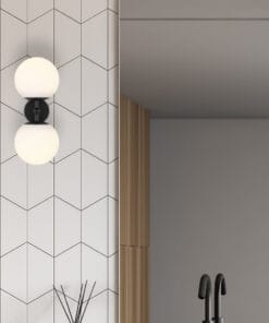 A modern bathroom features a geometric white tile wall, rectangular mirror, black faucet, Etern2 Wall Light with its double globe design, and a reed diffuser on the counter.