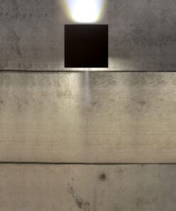 The Cube Wall Light casts white light beams up and down onto a smooth concrete wall, producing a striking visual effect.