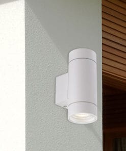 The KMAN Exterior Wall Light, a white cylindrical fixture, is installed on a light-colored exterior wall beside a wooden window frame.