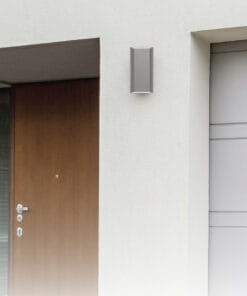 A modern exterior wall featuring a wooden front door, the Bloc LED Up-Down Wall Light in silver, and a sleek gray garage door.